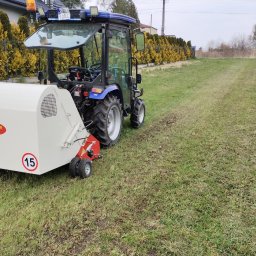 MarKop MARCIN KUCZMIERCZYK - Niebieski traktor z białym aeratorem na trawniku po aeracji. Widoczne ślady po przejeździe maszyny. W tle żywopłot i dom.