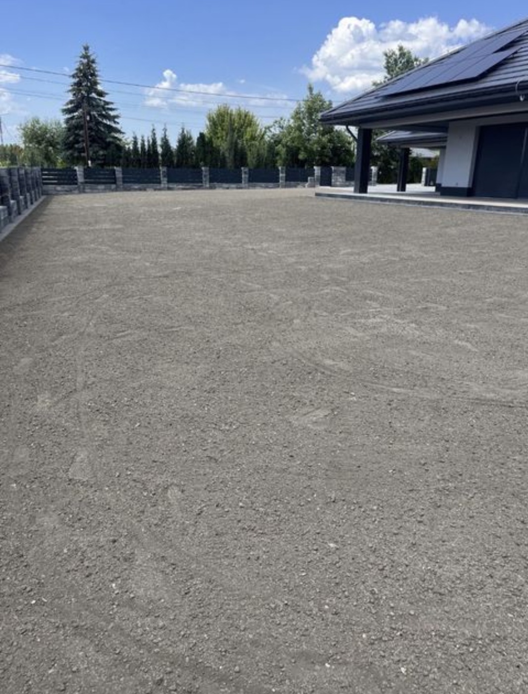 Przygotowany teren pod trawnik z widocznymi śladami opon, ogrodzony niskim, kamiennym płotem, z nowoczesnym domem w tle pod błękitnym niebem.