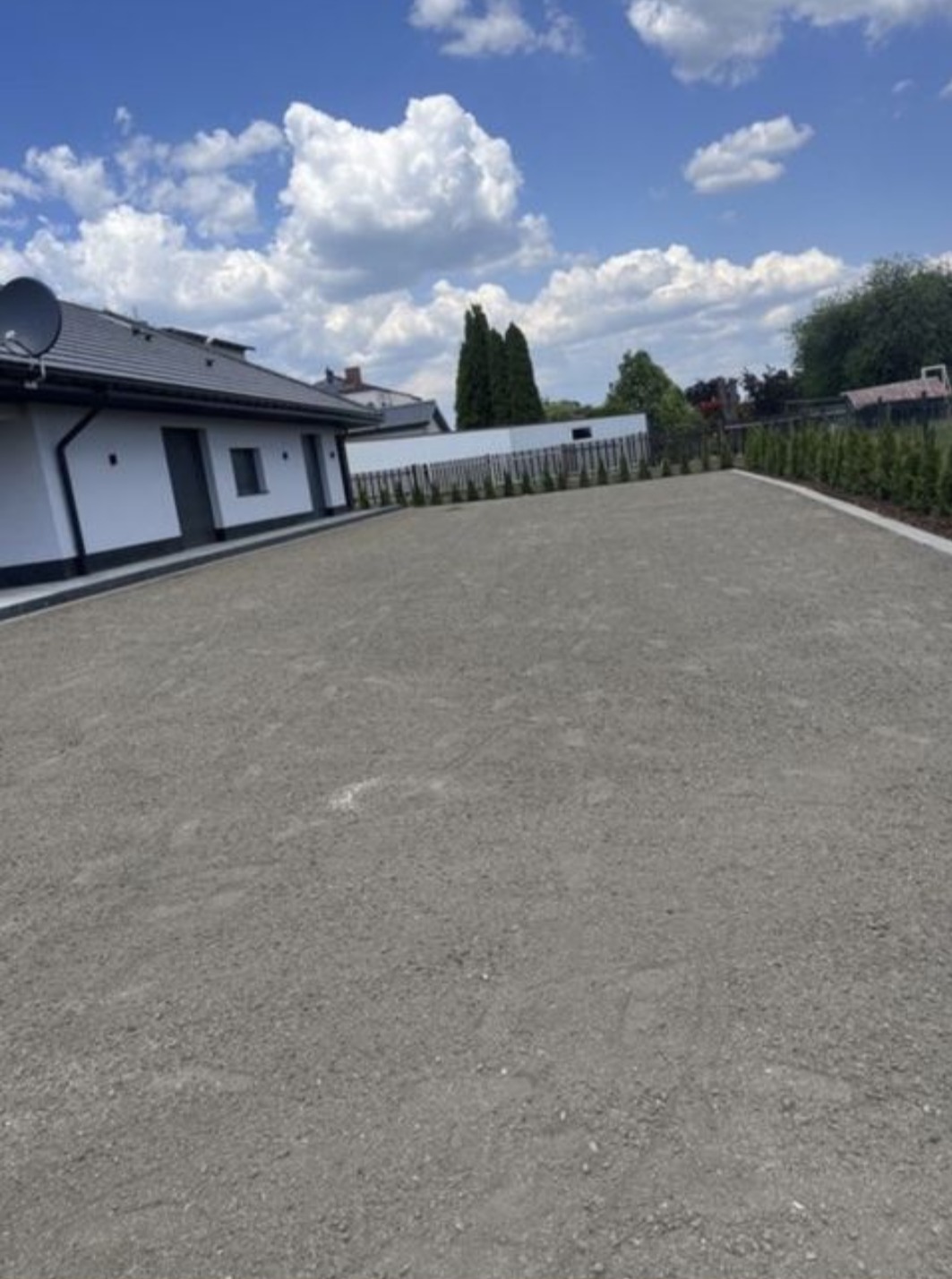 Utwardzony teren przed domem jednorodzinnym, gotowy pod dalsze prace. Widoczne obrzeża i nasadzenia z tui. Błękitne niebo z białymi chmurami w tle.