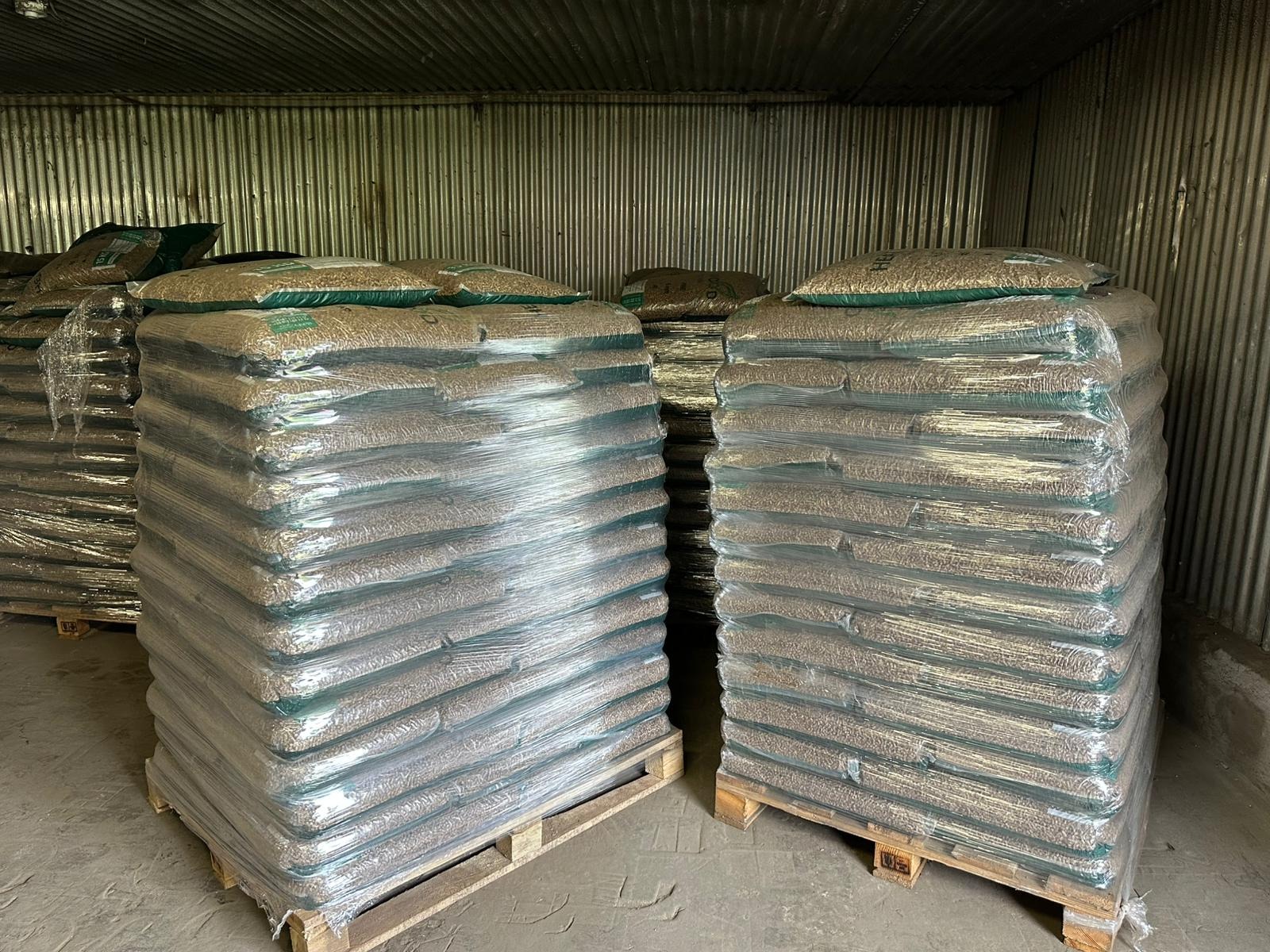 Stosy pelletu drzewnego na paletach, zabezpieczone folią, w magazynie o ścianach z blachy falistej. Widoczne worki z pelletem na górze stosów.