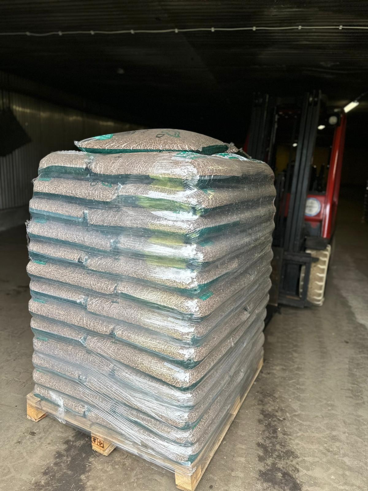 Stos worków z pelletem na palecie, owinięty folią, w magazynie w Urzucie. W tle wózek widłowy i blaszana ściana.