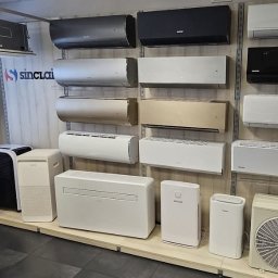 TERMO MOCNI - Ekspozycja różnorodnych klimatyzatorów Sinclair: ścienne, przenośne i kanałowe, w odcieniach bieli, szarości i czerni, prezentowane na tle ściany w showroomie.