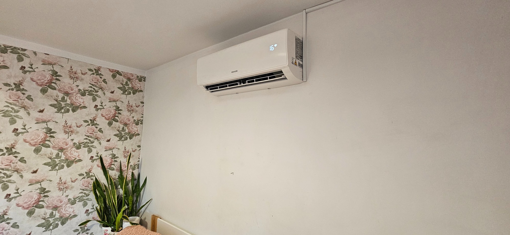 Biała klimatyzacja na ścianie w pokoju z tapetą w kwiaty i rośliną doniczkową. Widoczny wyświetlacz temperatury 16 stopni.