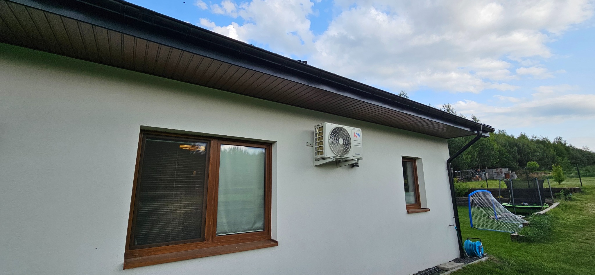 Boczny widok na biały budynek z zamontowaną klimatyzacją na ścianie, widoczne okna z brązowymi ramami i fragment ogrodu z zieloną trawą w tle.
