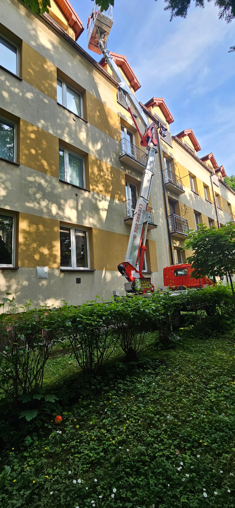 Podnośnik koszowy przy budynku mieszkalnym, prawdopodobnie podczas montażu instalacji. Widoczne balkony i zieleń.