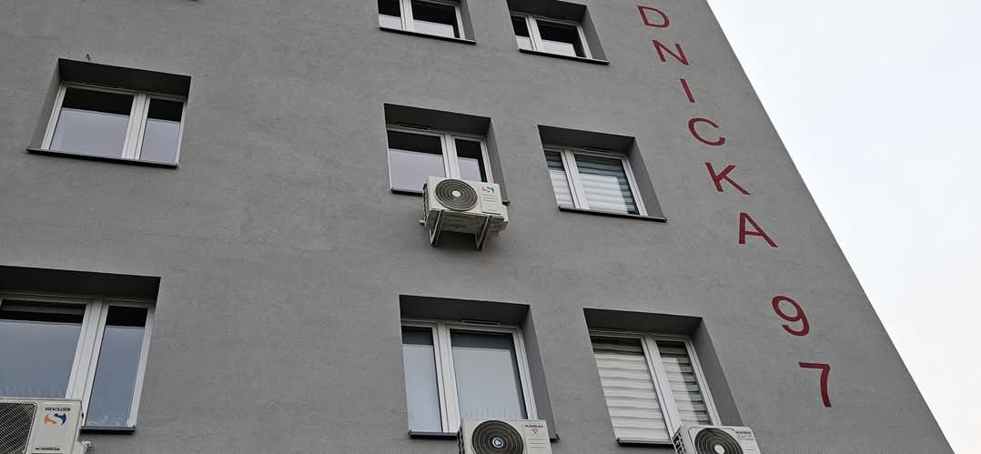Szara fasada budynku z oknami i zamontowanymi klimatyzatorami. Na ścianie pionowy napis 'DZNICKA 97' w kolorze czerwonym. Widok z dołu, pochmurne niebo.