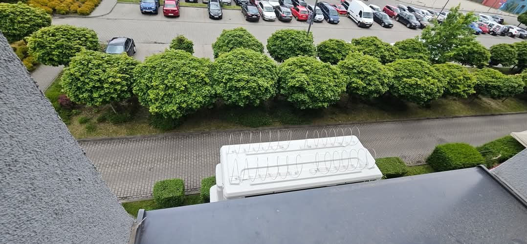 Biała klimatyzacja z metalowymi zabezpieczeniami na dachu budynku, widok z góry na parking i zieleń.