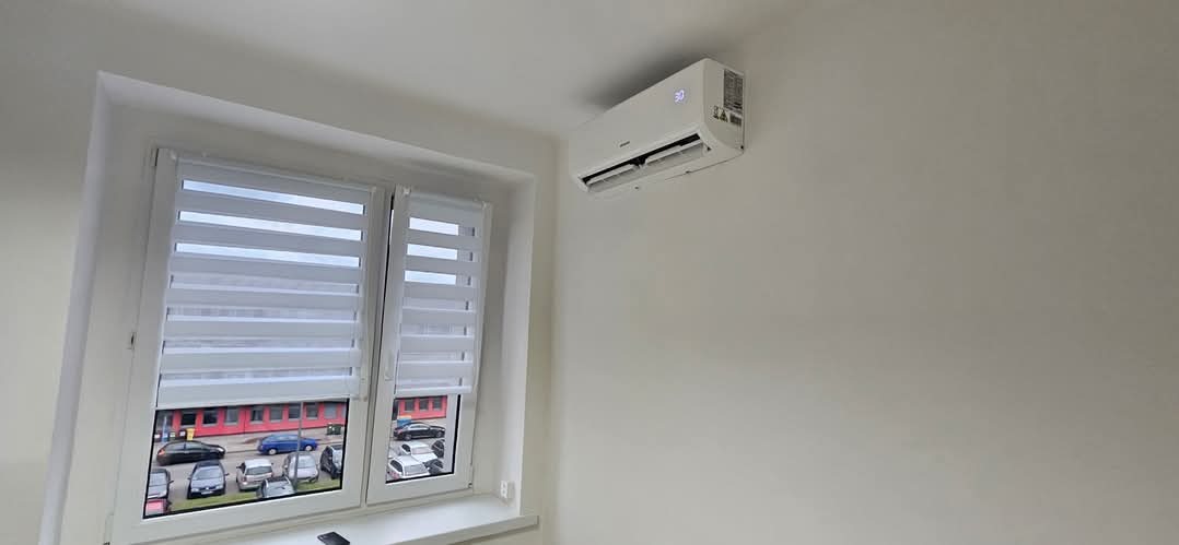 Biała klimatyzacja ścienna zamontowana w jasnym pokoju, widok na okno z żaluzjami i parking na zewnątrz. Minimalistyczny design.
