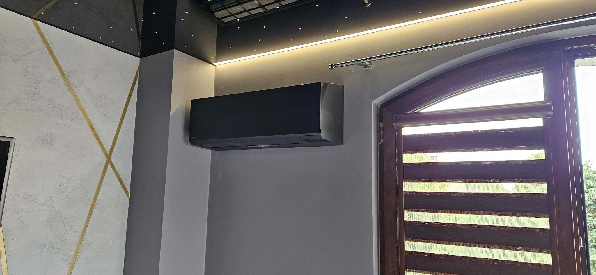 Czarny klimatyzator Toshiba na szarej ścianie w nowoczesnym wnętrzu z oknem i dekoracyjnym oświetleniem sufitowym.