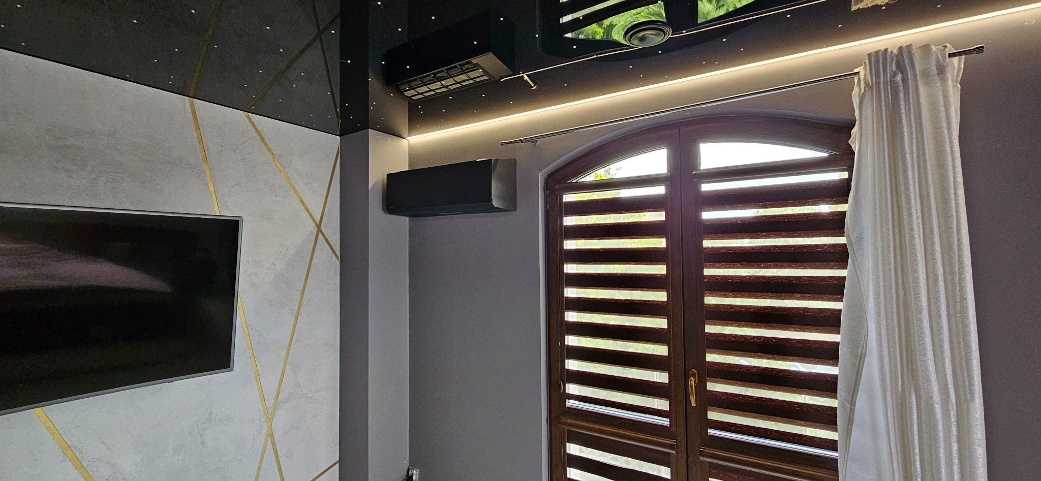 Nowoczesne wnętrze z klimatyzacją: czarny split system na szarej ścianie, dekoracyjny sufit z gwiazdami, okno z żaluzjami i telewizor na ścianie w geometryczne wzory.