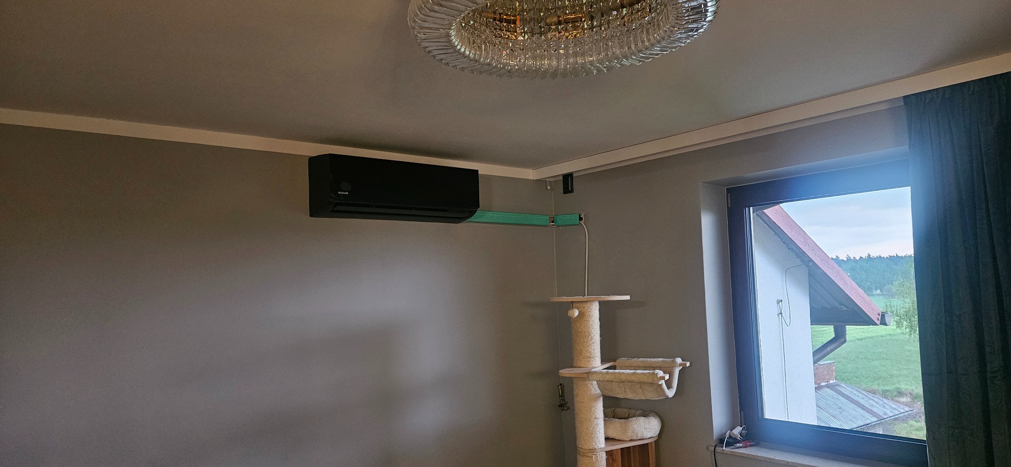 Czarny klimatyzator ścienny z nietypowym, jasnoniebieskim kanałem kablowym w pokoju z drapakiem dla kota i widokiem na zewnątrz przez okno.