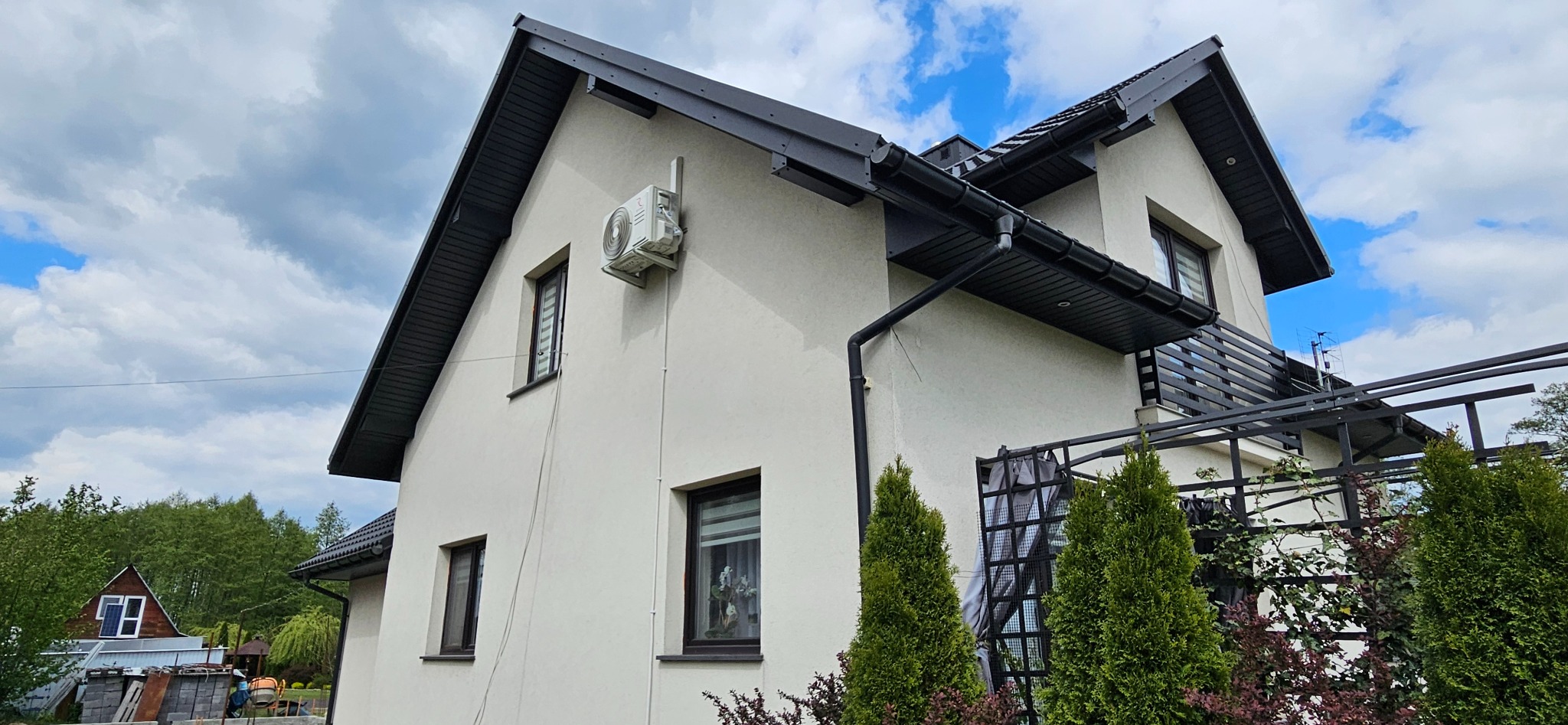 Biały dom jednorodzinny z zamontowaną klimatyzacją na bocznej ścianie. Widoczne okna z roletami i czarne rynny. Na pierwszym planie roślinność i elementy dekoracyjne.