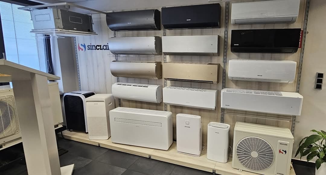 Ekspozycja różnorodnych klimatyzatorów Sinclair: ścienne, przenośne i kanałowe, w odcieniach bieli, szarości i czerni, prezentowane na tle ściany w showroomie.