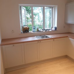 Philip Campbell - Jasna kuchnia z drewnianym blatem, widok na okno z narzędziami. Szafki z frezowanymi frontami i srebrnymi uchwytami. Na blacie bateria DeWalt.