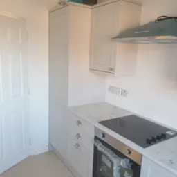 Jasna kuchnia z marmurowym blatem i płytą indukcyjną, okap w folii ochronnej. Widoczne szafki z szufladami i piekarnik. Nowoczesne wykończenie, minimalistyczny design.