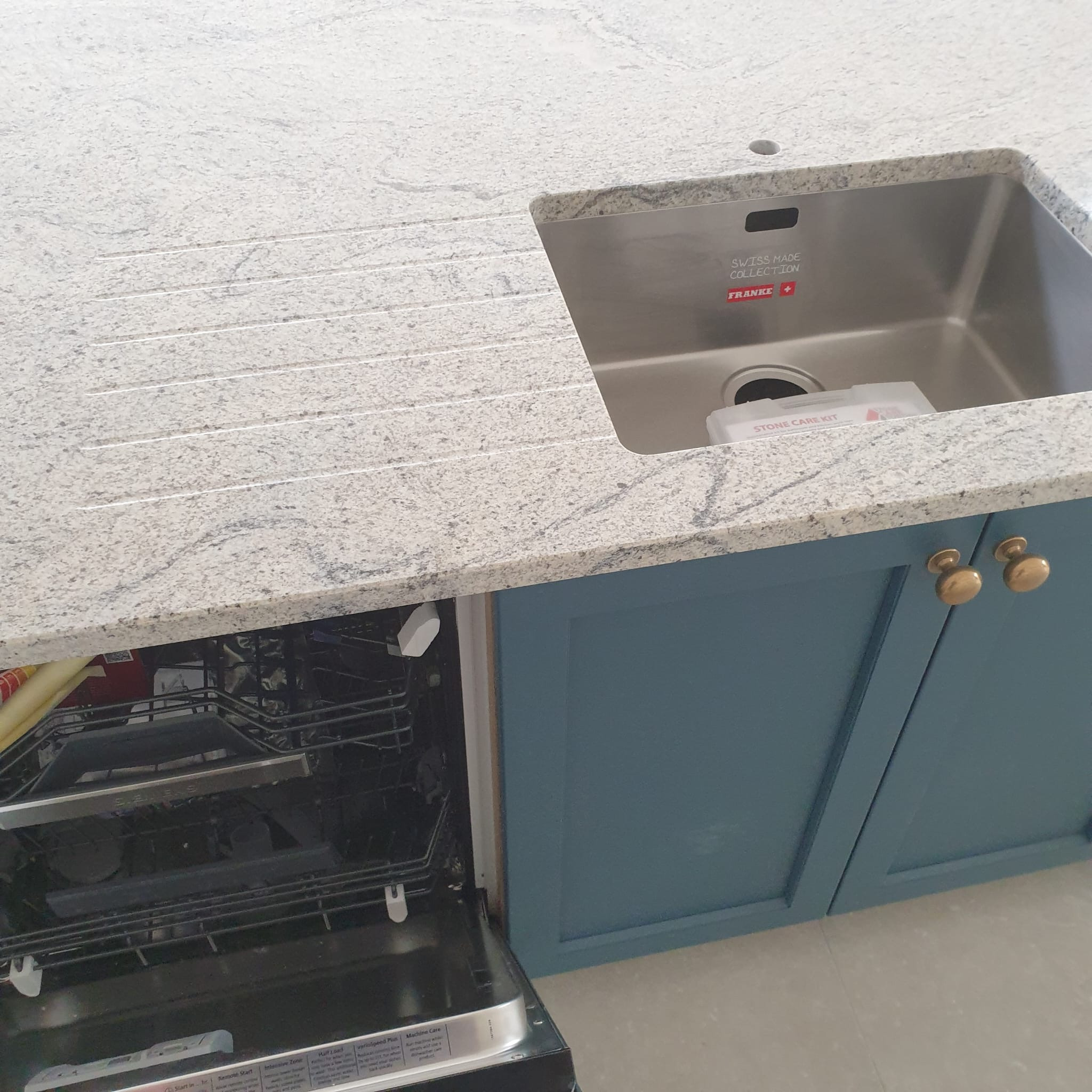 Granitowy blat kuchenny z wbudowanym zlewem Franke i otwartą zmywarką. Niebieskie szafki z złotymi uchwytami. Widoczny zestaw do pielęgnacji kamienia.