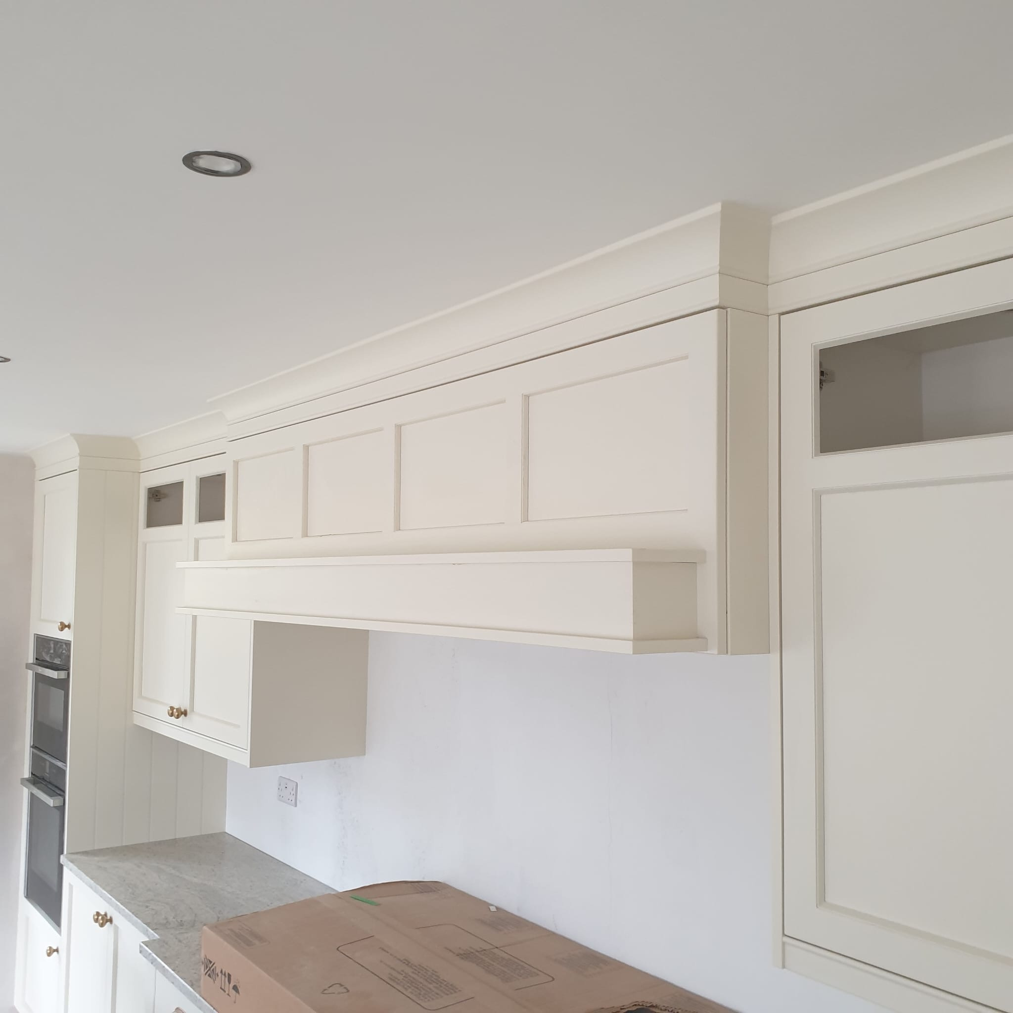 Elegancka, kremowa zabudowa kuchenna z frezowanymi frontami i szklanymi witrynami. Widoczny okap i fragment blatu. Całość w jasnym wnętrzu.