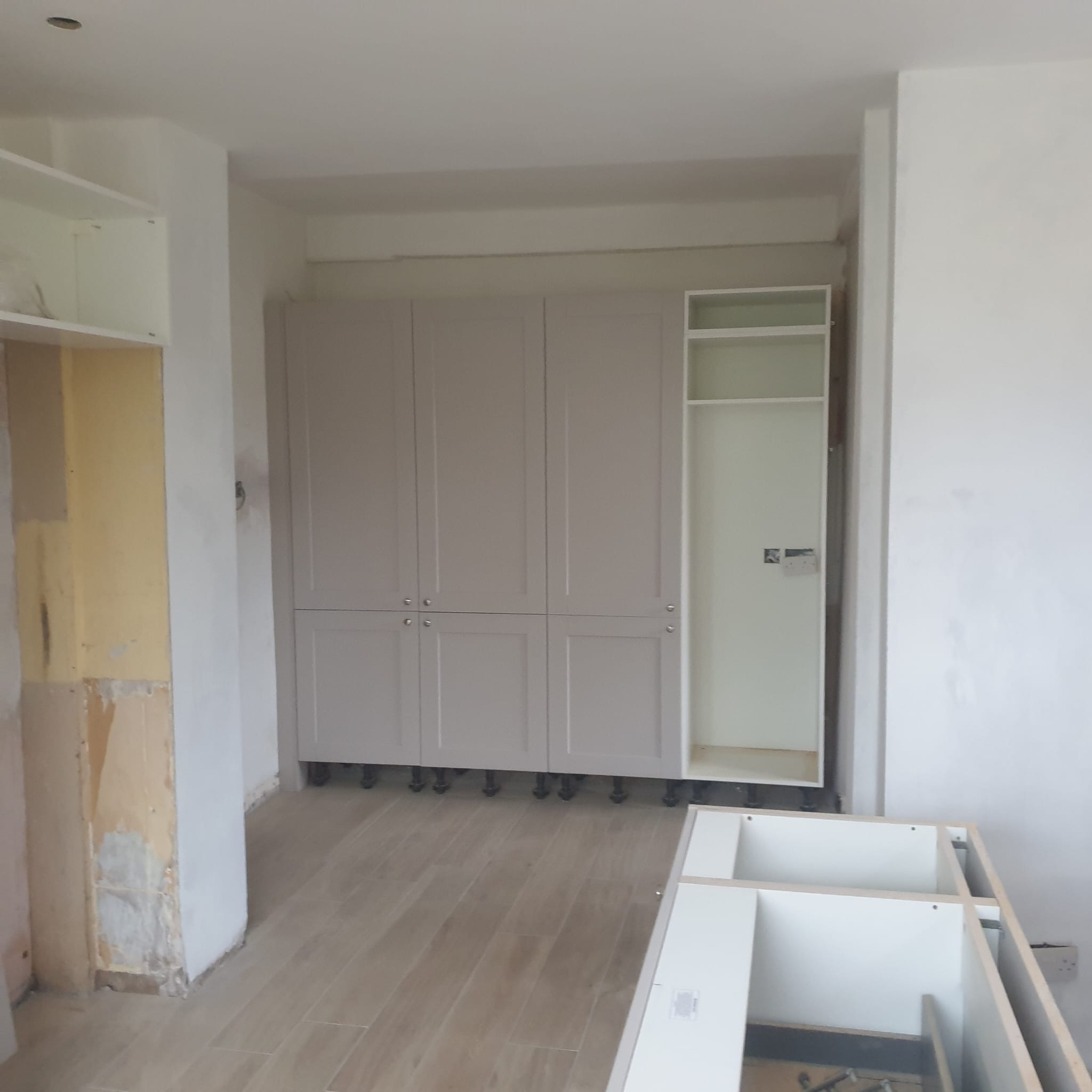 Montaż szafek kuchennych z szarymi frontami na regulowanych nóżkach. Widoczne elementy w trakcie instalacji, surowa ściana i drewnopodobna podłoga.