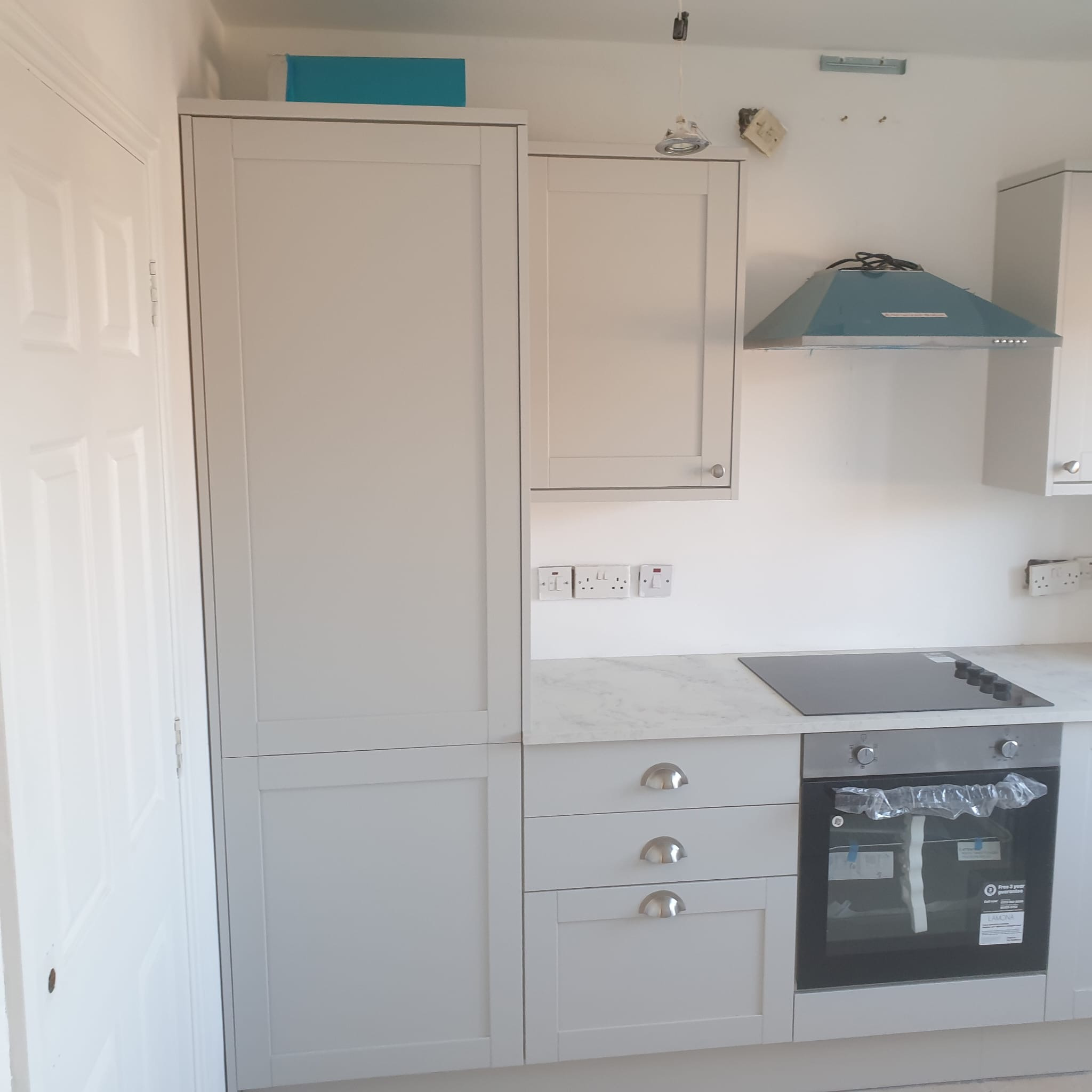 Jasna kuchnia z zabudową: szafki wiszące i stojące z szufladami, płyta indukcyjna na blacie. Nowy piekarnik zabezpieczony folią. Świeżo zamontowany okap.