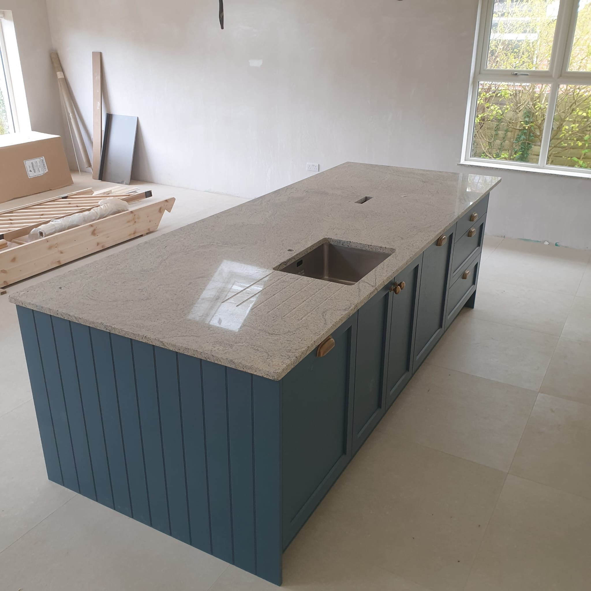 Wyspa kuchenna z szarym blatem i niebieskimi frontami szafek, z wbudowanym zlewem. W tle widoczne materiały budowlane i okno, sugerujące remont kuchni.
