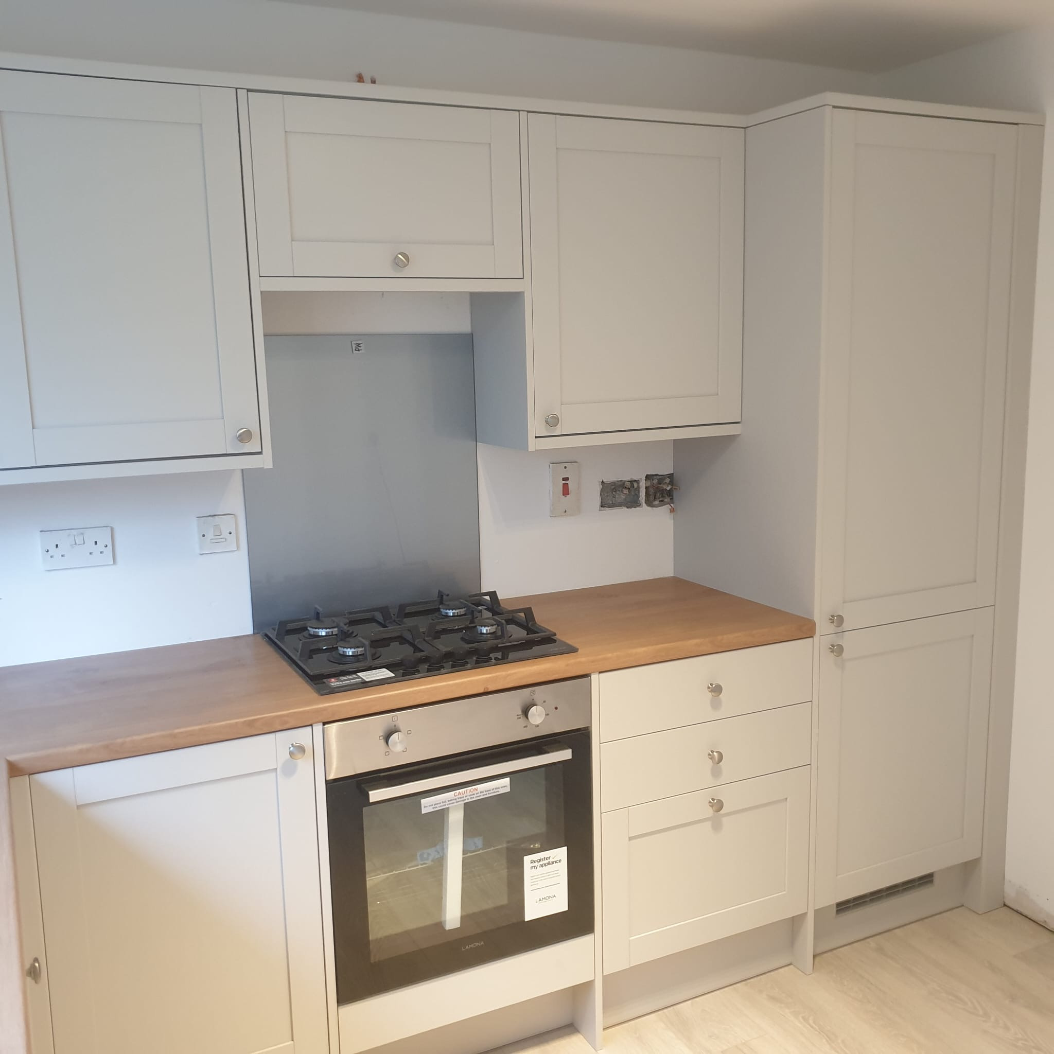 Jasnoszara zabudowa kuchenna z drewnianym blatem, płyta gazowa i piekarnik marki Lamona. Widoczne gniazdka elektryczne i niedokończone wykończenie ściany.