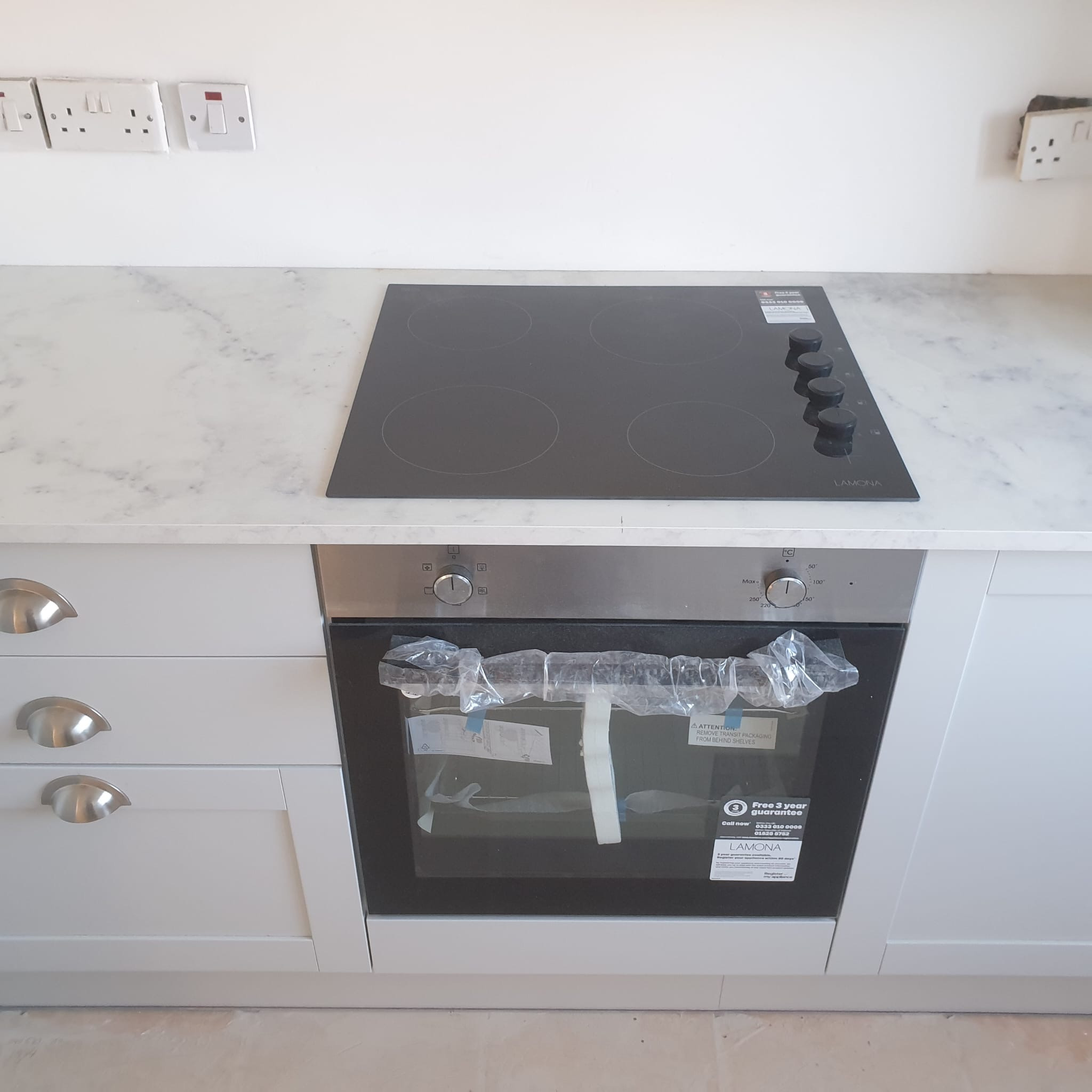 Nowa płyta indukcyjna Lamona i piekarnik zamontowane w białej zabudowie kuchennej z marmurowym blatem. Piekarnik jeszcze w folii ochronnej.