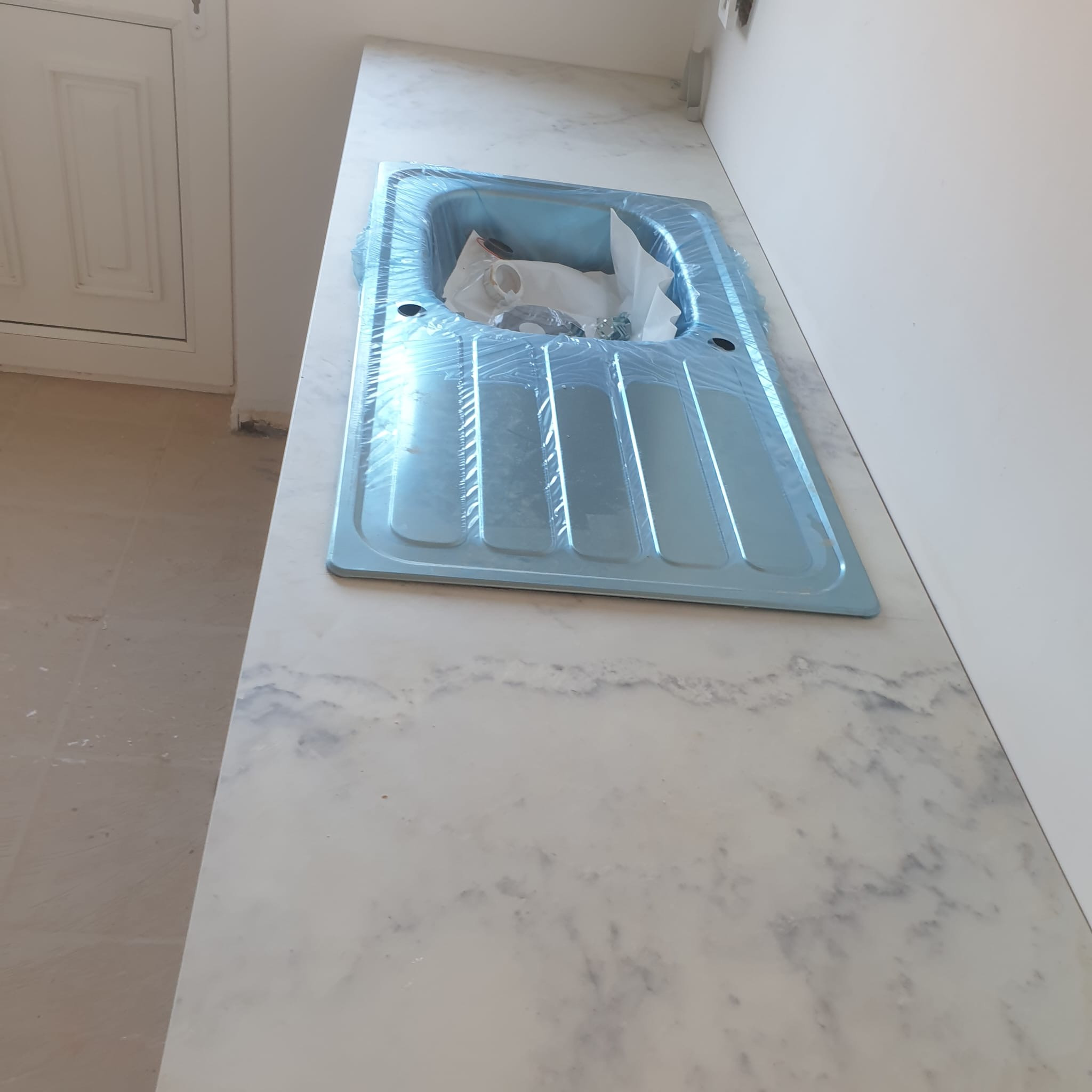 Błękitny zlew kuchenny, zabezpieczony folią, na blacie z marmurowym wzorem, po montażu w nowej kuchni. Widoczny fragment podłogi w trakcie prac wykończeniowych.