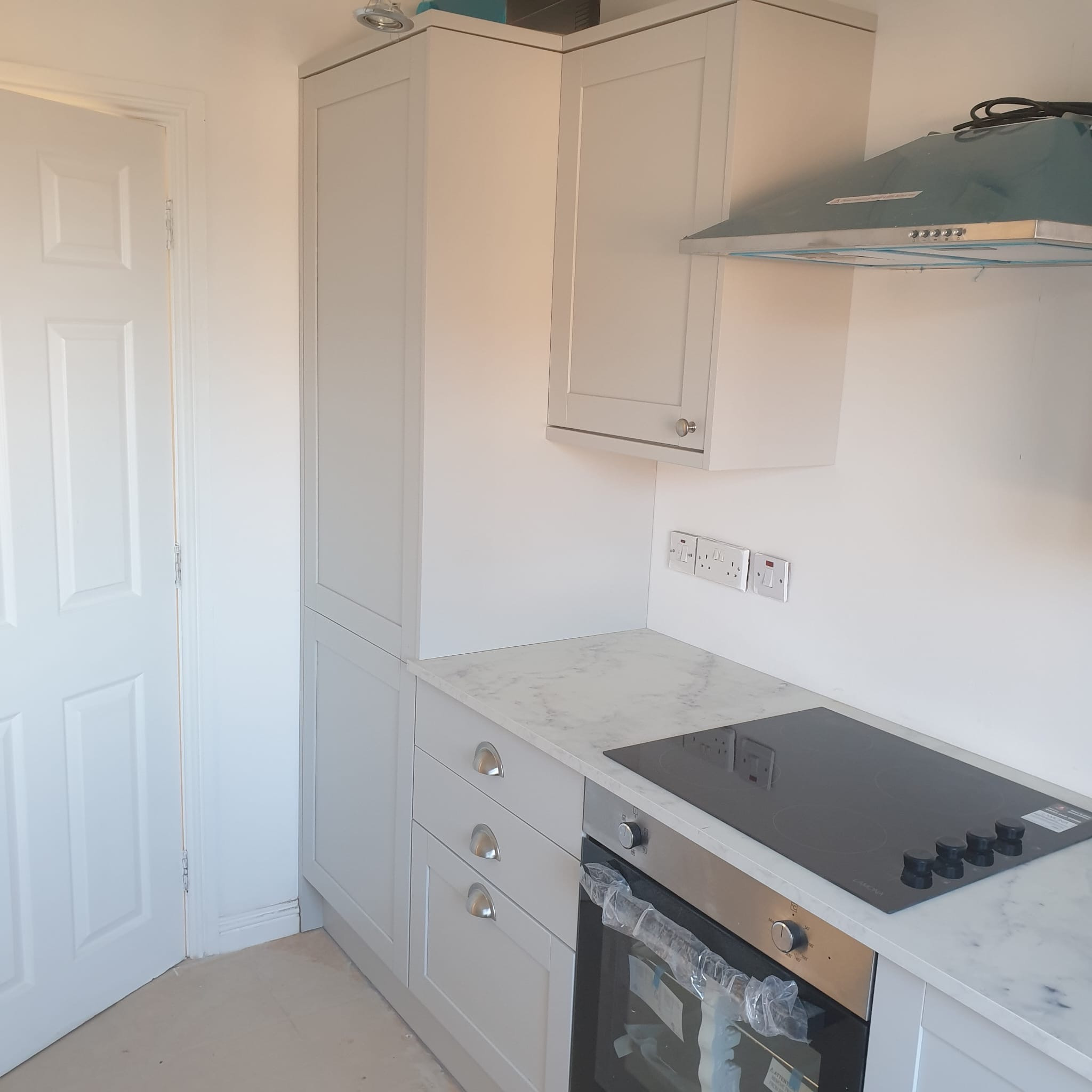 Jasna kuchnia z marmurowym blatem i płytą indukcyjną, okap w folii ochronnej. Widoczne szafki z szufladami i piekarnik. Nowoczesne wykończenie, minimalistyczny design.