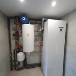 Biały piec York z zbiornikiem i niebieskim naczyniem wzbiorczym, połączony rurami w pomieszczeniu wykończonym płytkami. Widoczne liczne zawory i pompy obiegowe.