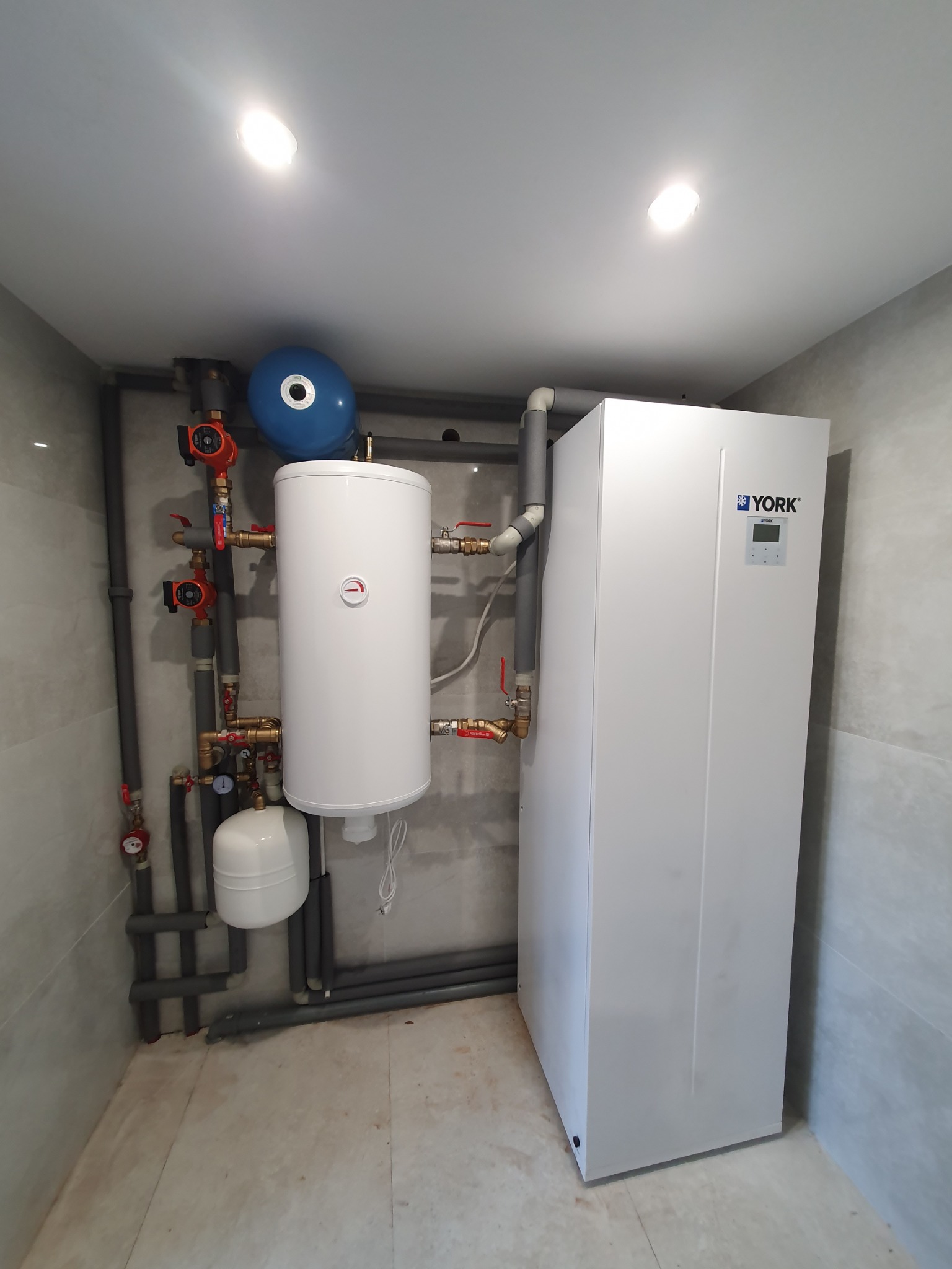Biały piec York z zbiornikiem i niebieskim naczyniem wzbiorczym, połączony rurami w pomieszczeniu wykończonym płytkami. Widoczne liczne zawory i pompy obiegowe.