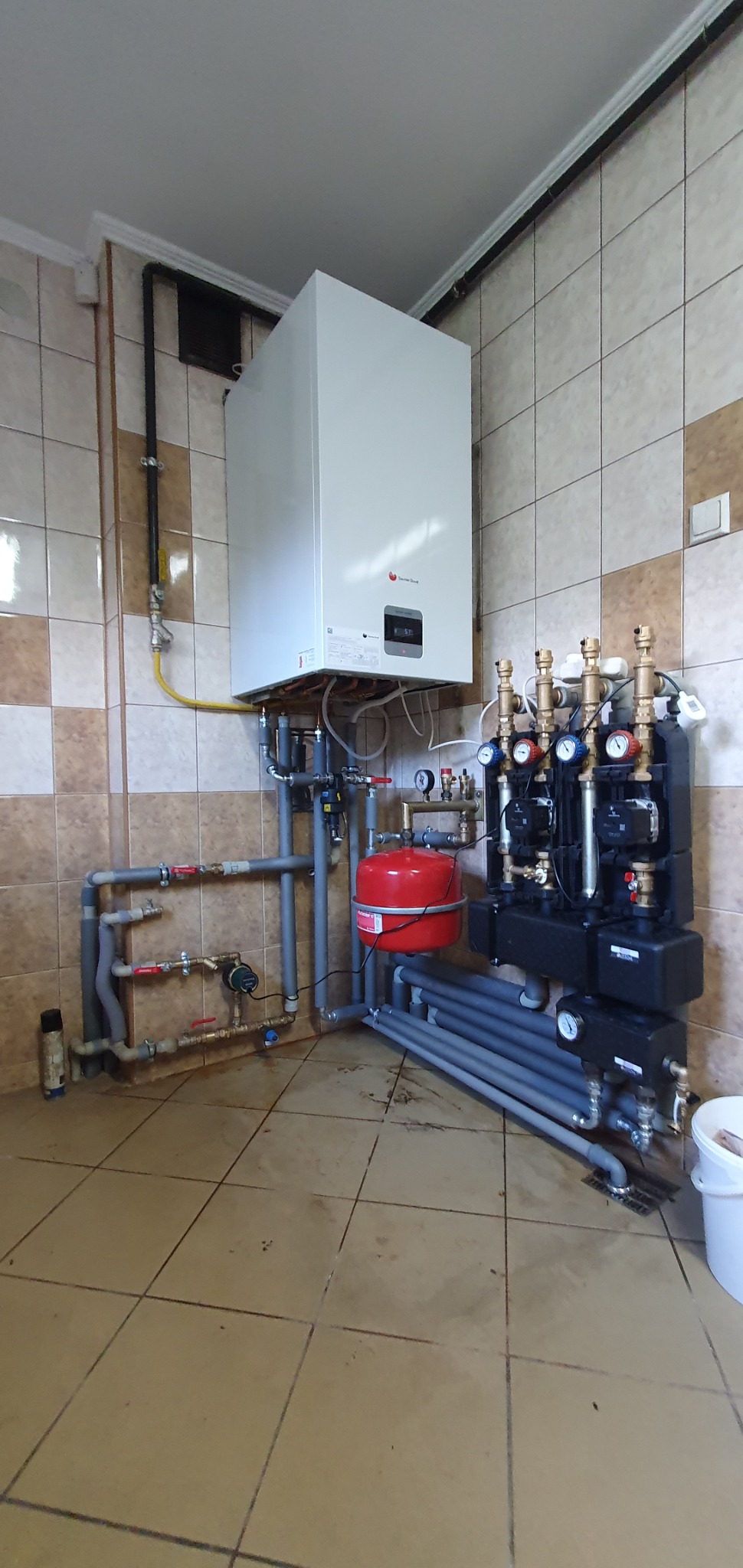 Złożona instalacja CO z piecem gazowym, zbiornikiem wyrównawczym i rozdzielaczem. Widoczne rury, zawory, manometry. Ściany wyłożone płytkami w odcieniach beżu i brązu.