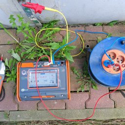 ENERGOSYSTEM PIOTR MIERNIK - Pomiar rezystancji uziemienia instalacji odgromowej miernikiem Sonel MPI-540-PV na tle ceglanej kostki brukowej i roślinności.