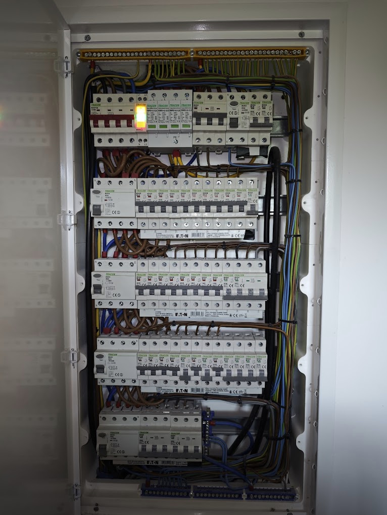 Rozdzielnica elektryczna w białej obudowie, widoczne liczne zabezpieczenia, przewody i listwy zaciskowe. Jeden z bezpieczników sygnalizuje stan aktywny.