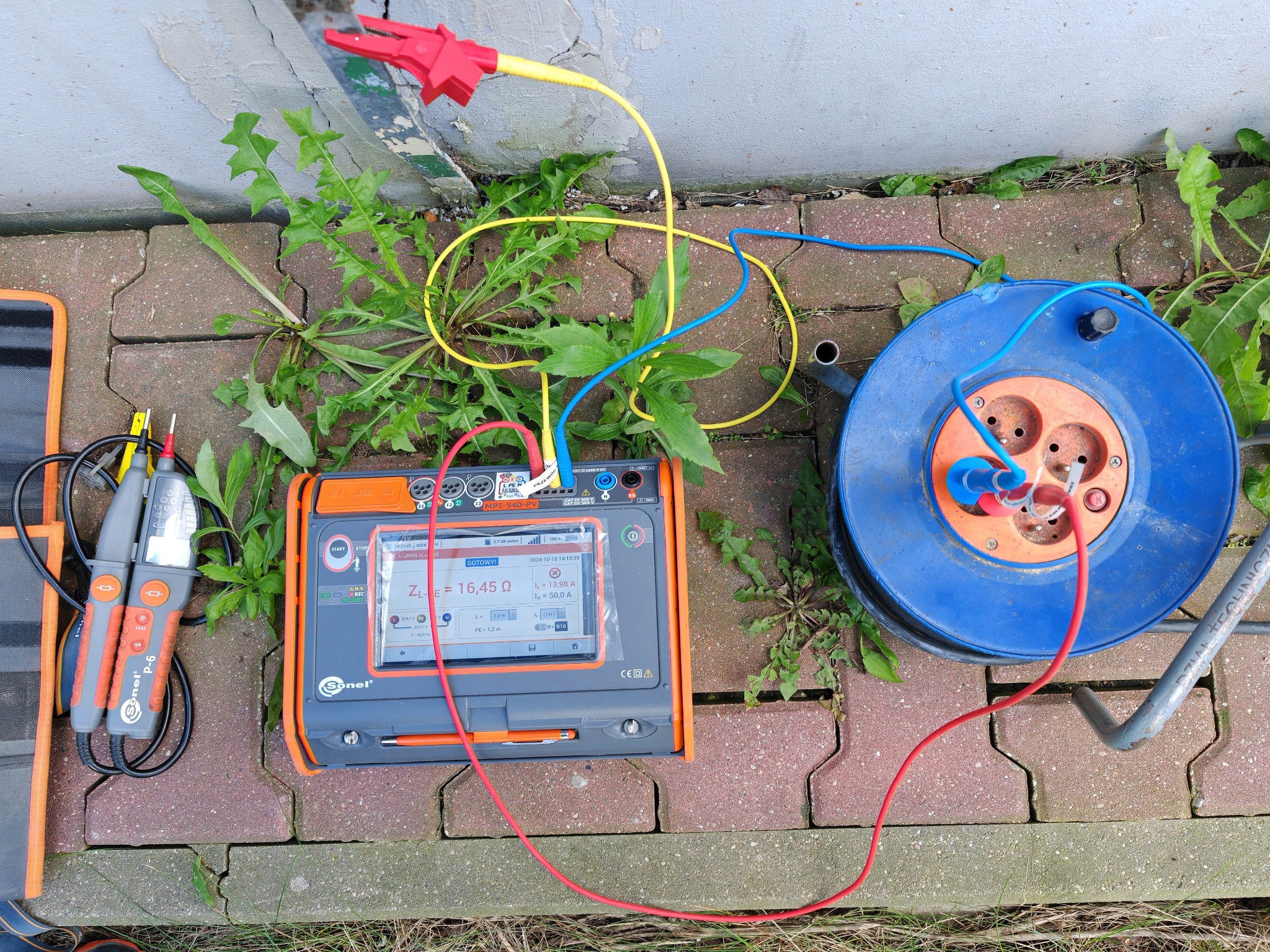 Pomiar rezystancji uziemienia instalacji odgromowej miernikiem Sonel MPI-540-PV na tle ceglanej kostki brukowej i roślinności.