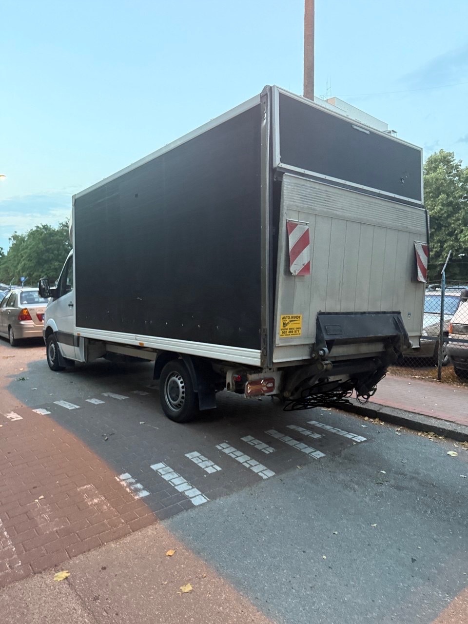 Tył białego busa dostawczego z czarną nadbudową zaparkowanego na ulicy z namalowanymi pasami dla pieszych, widok z perspektywy chodnika.