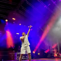 Wokalista w białym stroju na scenie, oświetlony reflektorami w kolorach fioletu i czerwieni, z uniesioną ręką i mikrofonem, w tle widoczny DJ.