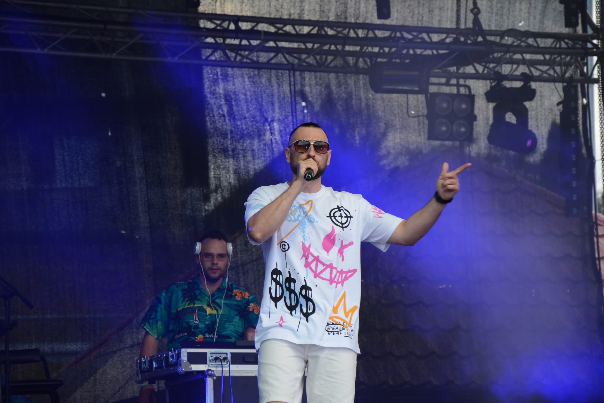 Występ zespołu muzycznego z Sitna: wokalista w okularach, biała koszulka z nadrukiem, DJ w tle, scena z oświetleniem, koncert plenerowy.