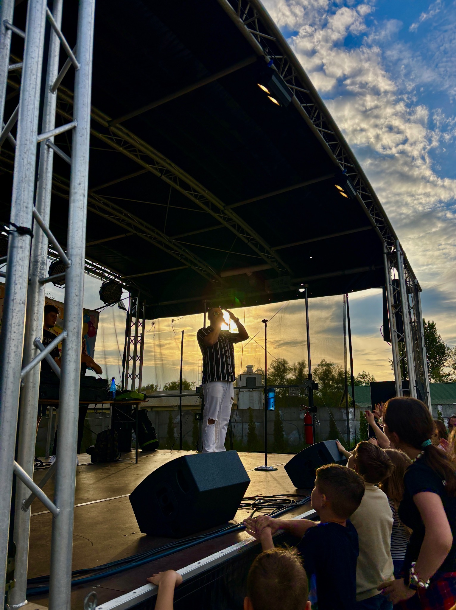 Wokalista w paski na scenie, śpiewa do mikrofonu, w tle zachodzące słońce i publiczność. Koncert plenerowy z widocznymi głośnikami i konstrukcją sceny.