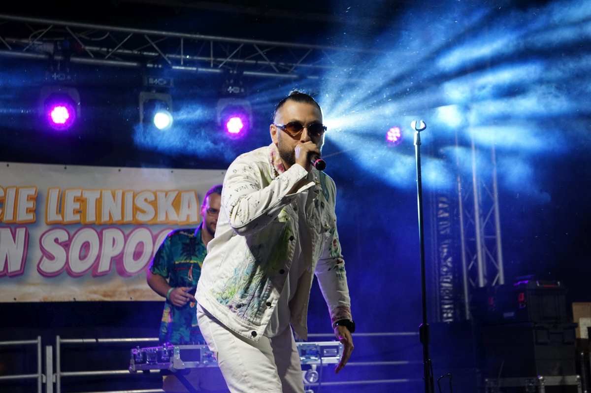 Wokalista w białym stroju i okularach śpiewa na scenie, w tle oświetlenie sceniczne i fragment napisu 'Letniska w Sopocie'.