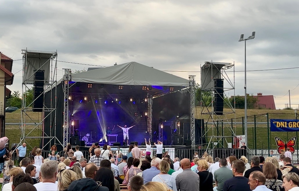 Plenerowy koncert zespołu muzycznego na scenie z oświetleniem, widownia siedząca na ławkach, w tle budynki i pochmurne niebo.