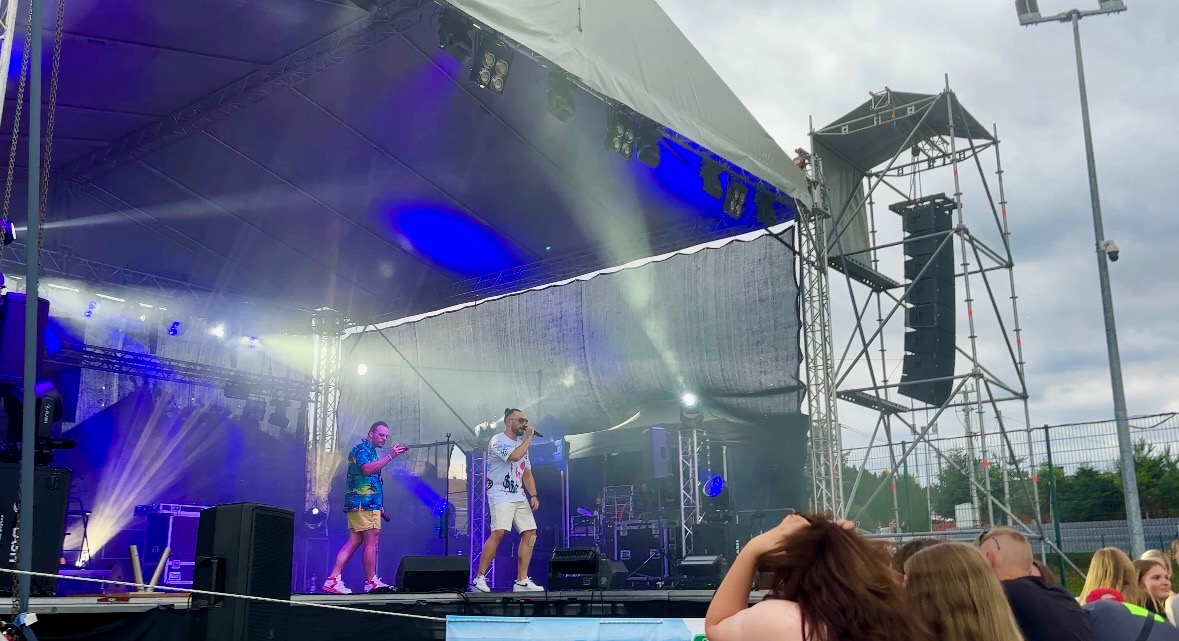 Zespół muzyczny na scenie podczas koncertu plenerowego, oświetlonej niebieskimi światłami, widoczna publiczność i konstrukcja nagłośnienia.