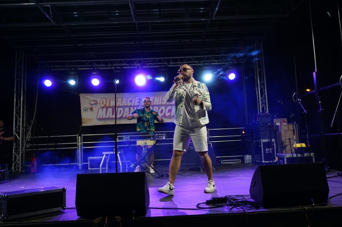 Wokalista w białym komplecie i okularach śpiewa na scenie z mikrofonem, za nim klawiszowiec i banner 'Otwarcie Majdan Rock'. Scena oświetlona reflektorami.