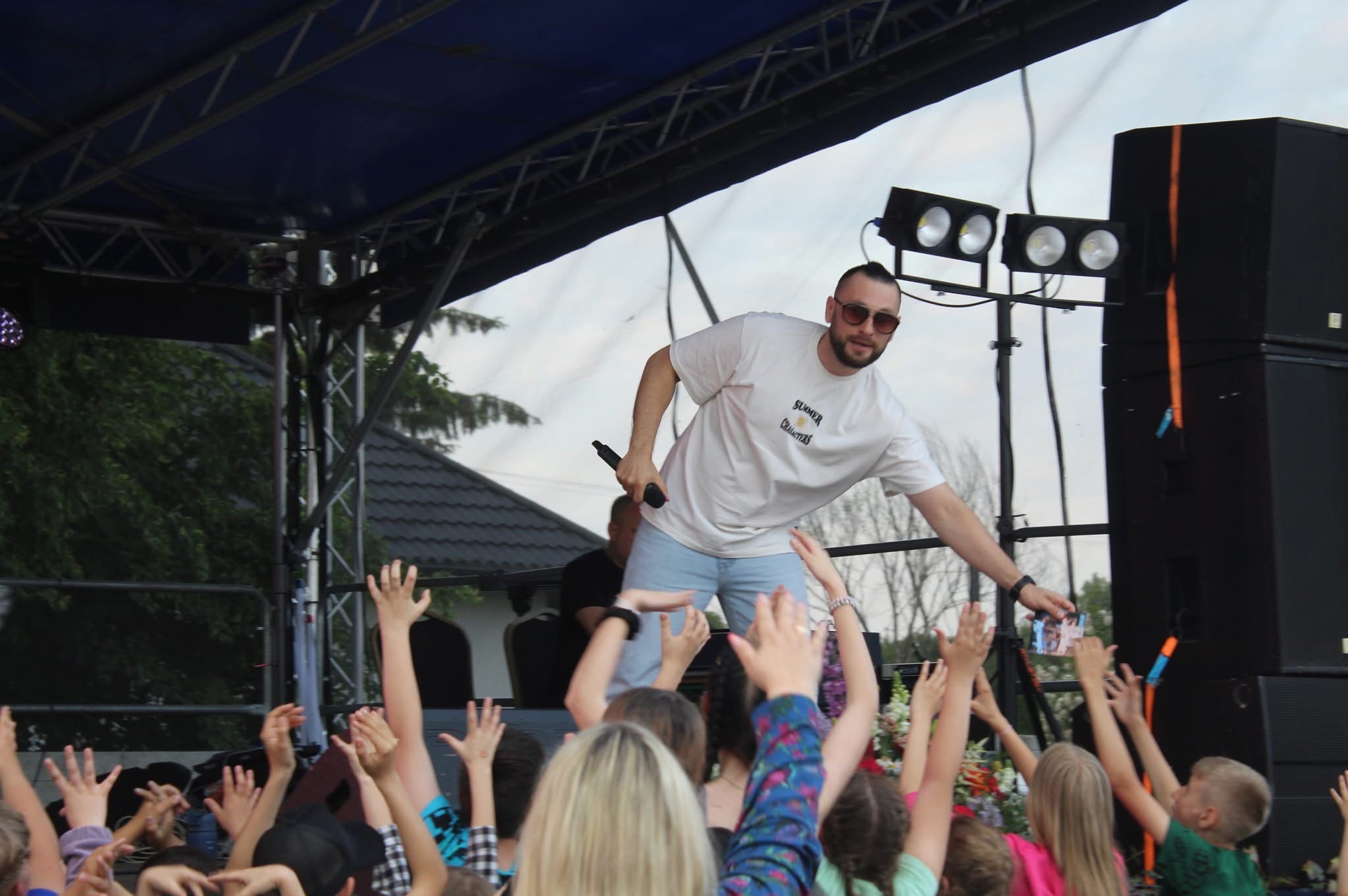 Występujący w okularach na scenie, z mikrofonem w dłoni, w otoczeniu rozentuzjazmowanego tłumu z podniesionymi rękami podczas koncertu.