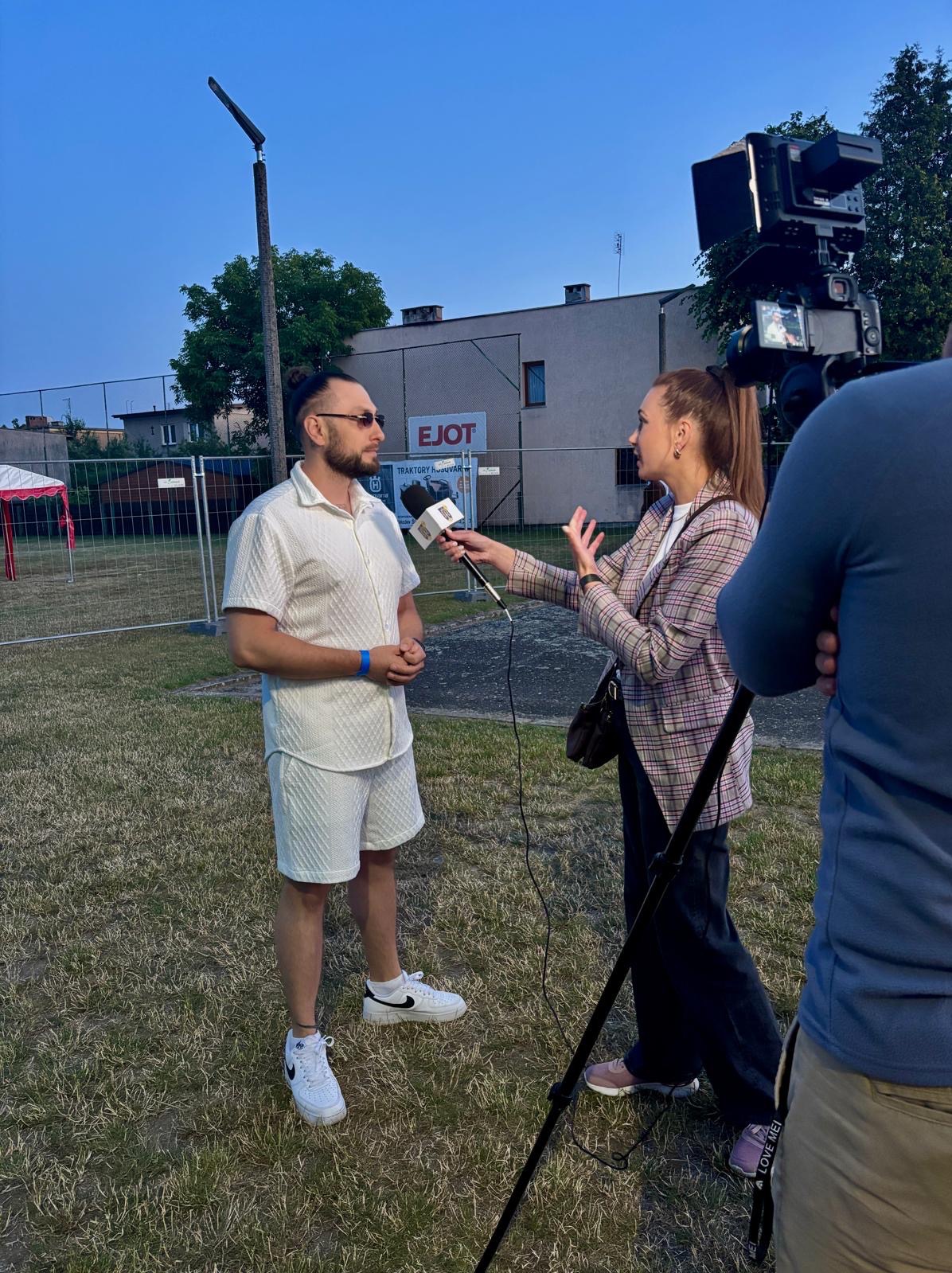 Wywiad z muzykiem w białym komplecie na tle sceny plenerowej. Reporterka z mikrofonem, kamera i operator w kadrze. Ujęcie z dołu, wieczorne oświetlenie.