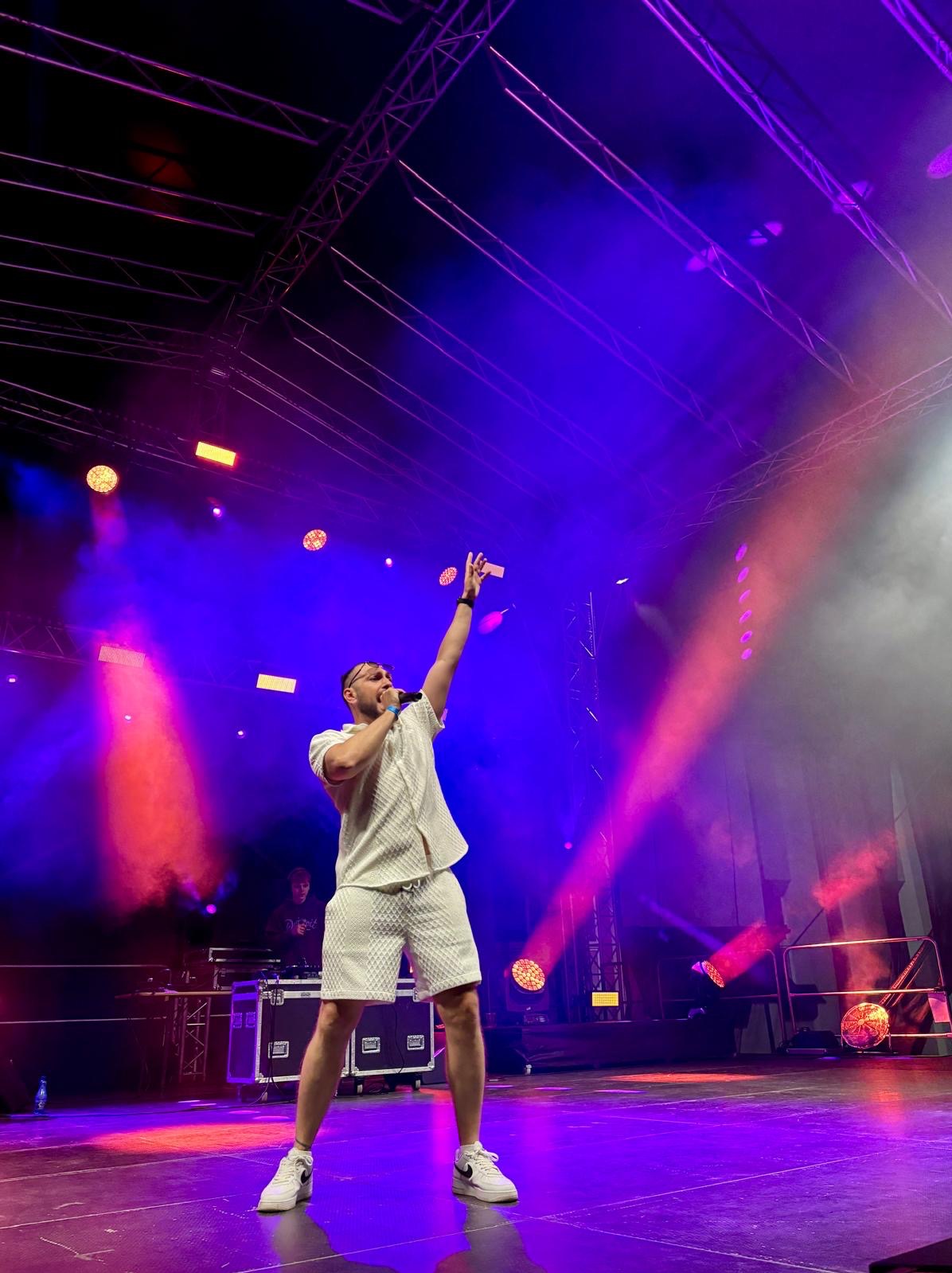 Wokalista w białym stroju na scenie, oświetlony reflektorami w kolorach fioletu i czerwieni, z uniesioną ręką i mikrofonem, w tle widoczny DJ.