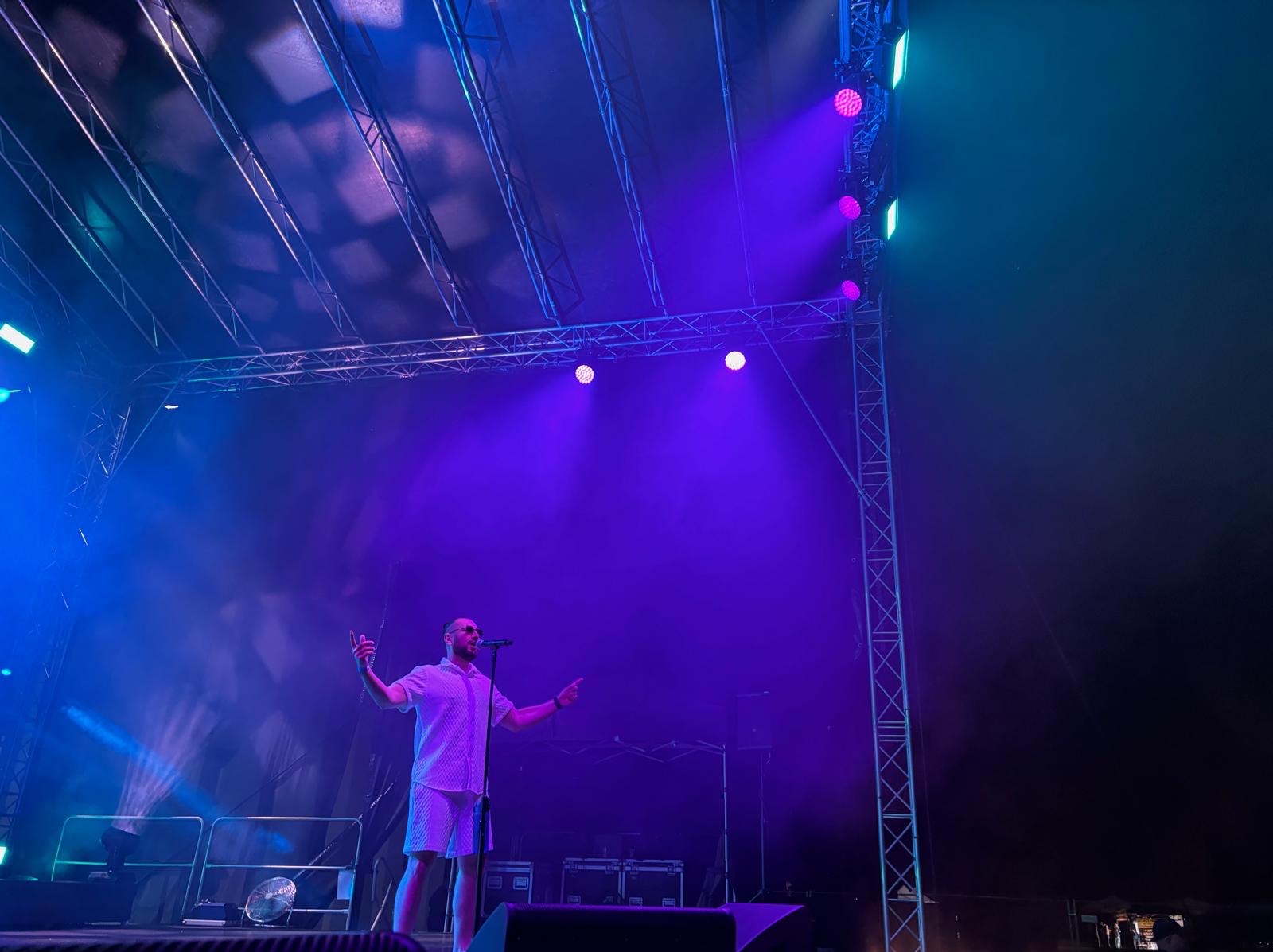 Wokalista w białym stroju na scenie oświetlonej niebiesko-fioletowymi światłami, z mikrofonem i konstrukcją metalową w tle podczas występu.
