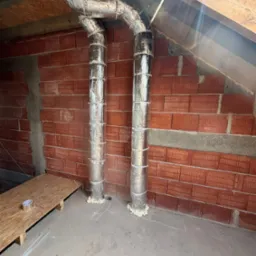 Dwa pionowe przewody wentylacyjne w izolacji, zamocowane do ściany z czerwonej cegły w surowym wnętrzu poddasza. Widoczne elementy konstrukcji dachu i podłogi.