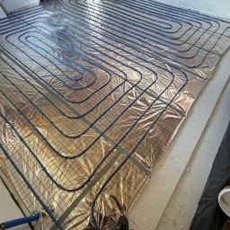 BartSystem Bartosz Kaczmarski - Rozłożona instalacja ogrzewania podłogowego na folii z siatką w surowym wnętrzu. Widoczne niebieskie rury ułożone w pętle i fragment osoby w kadrze.