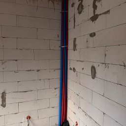 BartSystem Bartosz Kaczmarski - Instalacja ogrzewania podłogowego w surowym wnętrzu: rury grzewcze na folii, pionowe rury wodne (czerwona i niebieska) przy ścianie z betonu komórkowego.