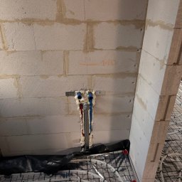 BartSystem Bartosz Kaczmarski - Instalacja ogrzewania podłogowego w trakcie montażu: rury grzewcze ułożone na izolacji, pionowe rury doprowadzające zabezpieczone pianką montażową przy ścianie z betonu komórkowego.